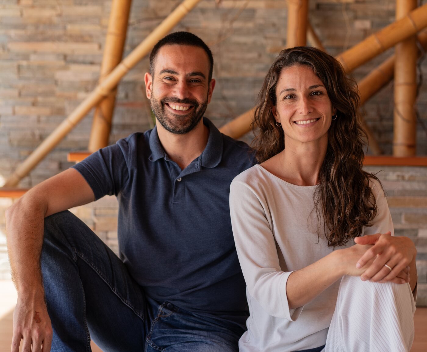 Sergi Colomer i Elisabeth del Río asseguts davant d’una paret de pedra i canyes de bambú, somrient a càmera